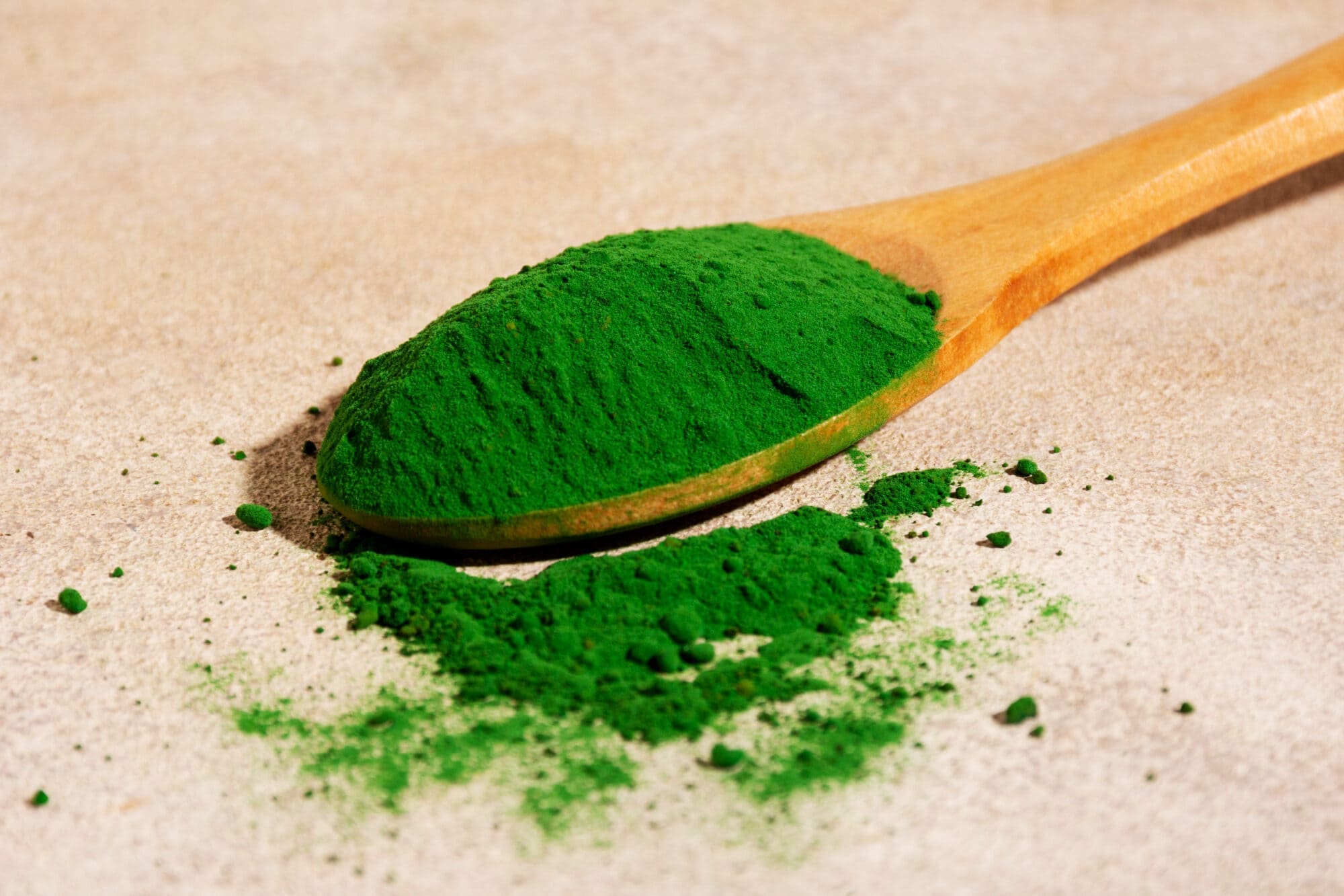 A close-up of vibrant green powder on a wooden spoon, with powder scattered on a textured beige surface.